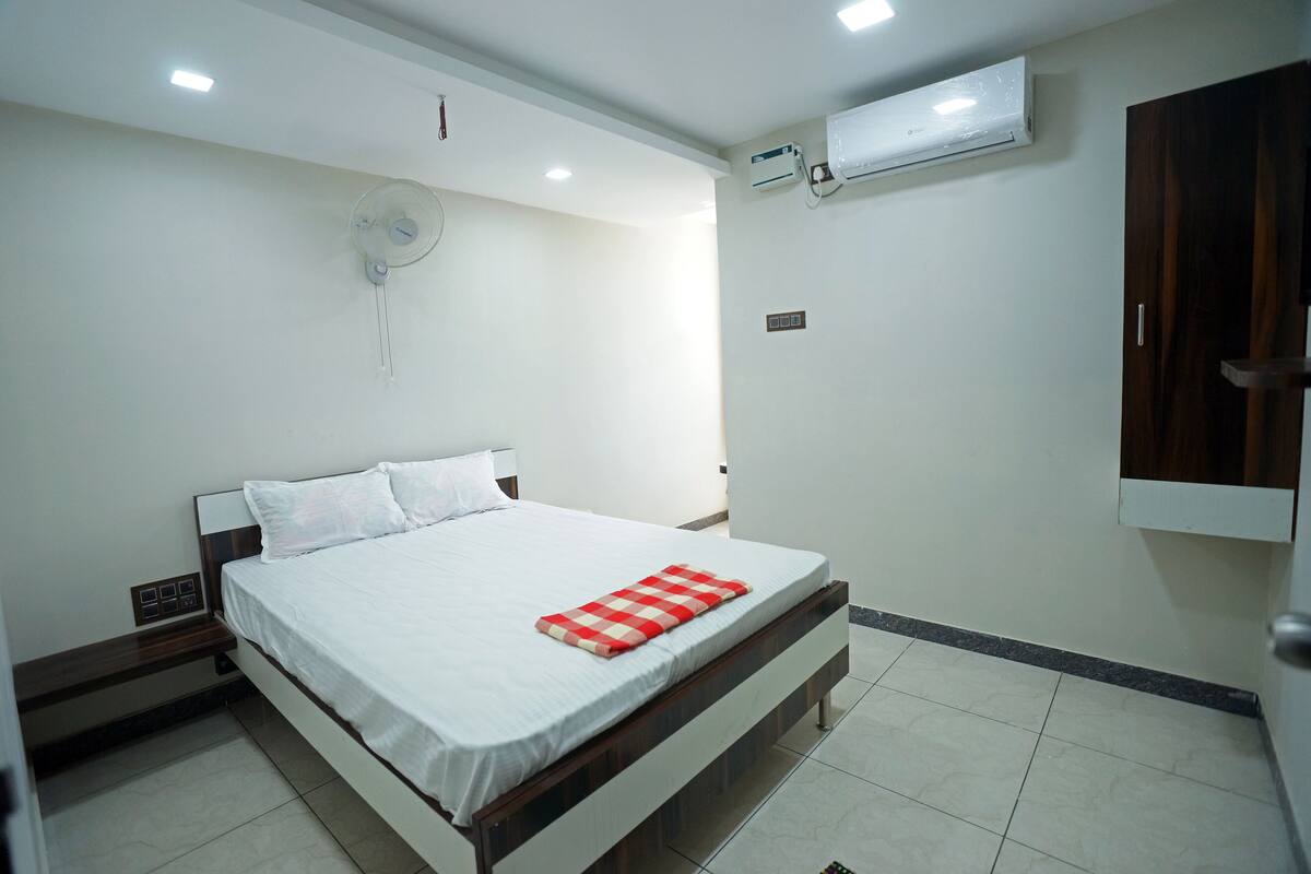 Ein ordentlich eingerichtetes Schlafzimmer mit Bett, Nachttisch, Wandventilator und Klimaanlage.