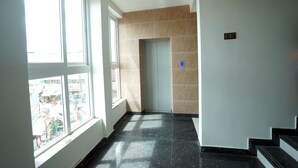 A hallway with a view of a busy street through large windows, a door at the end, and a staircase on the right. - Sai Lodge (Tiruchirappalli)