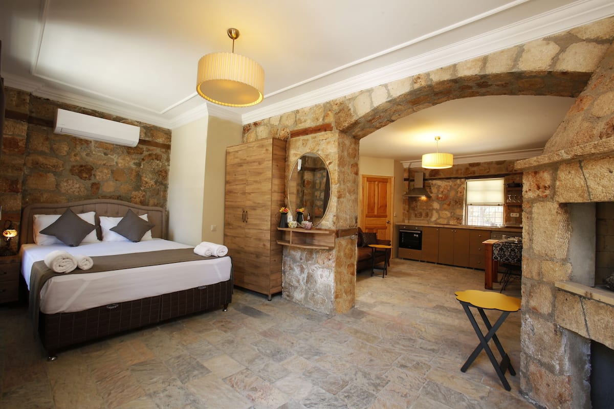 A bedroom with a stone wall, a wooden bed, a small table, and a kitchen area with a stone fireplace.