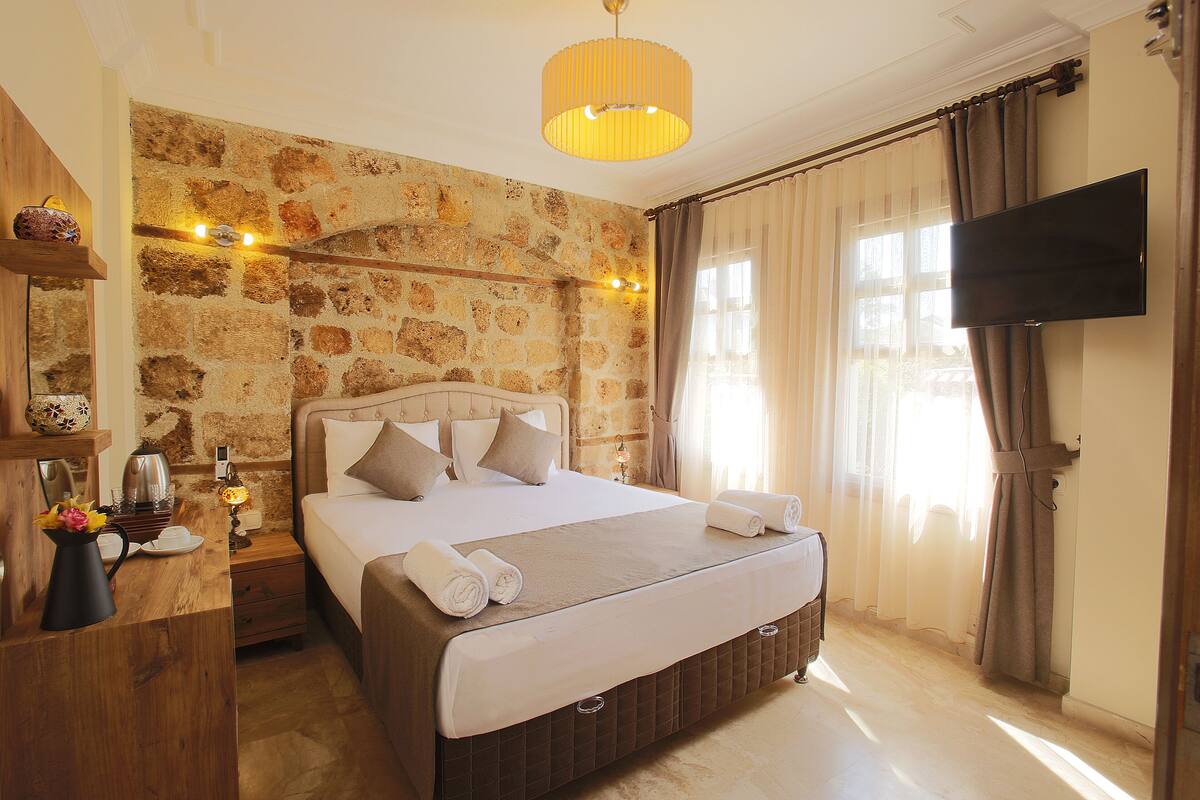 A bedroom with a stone wall, a bed with a headboard, a bedside table with a lamp, and a TV mounted on the wall.