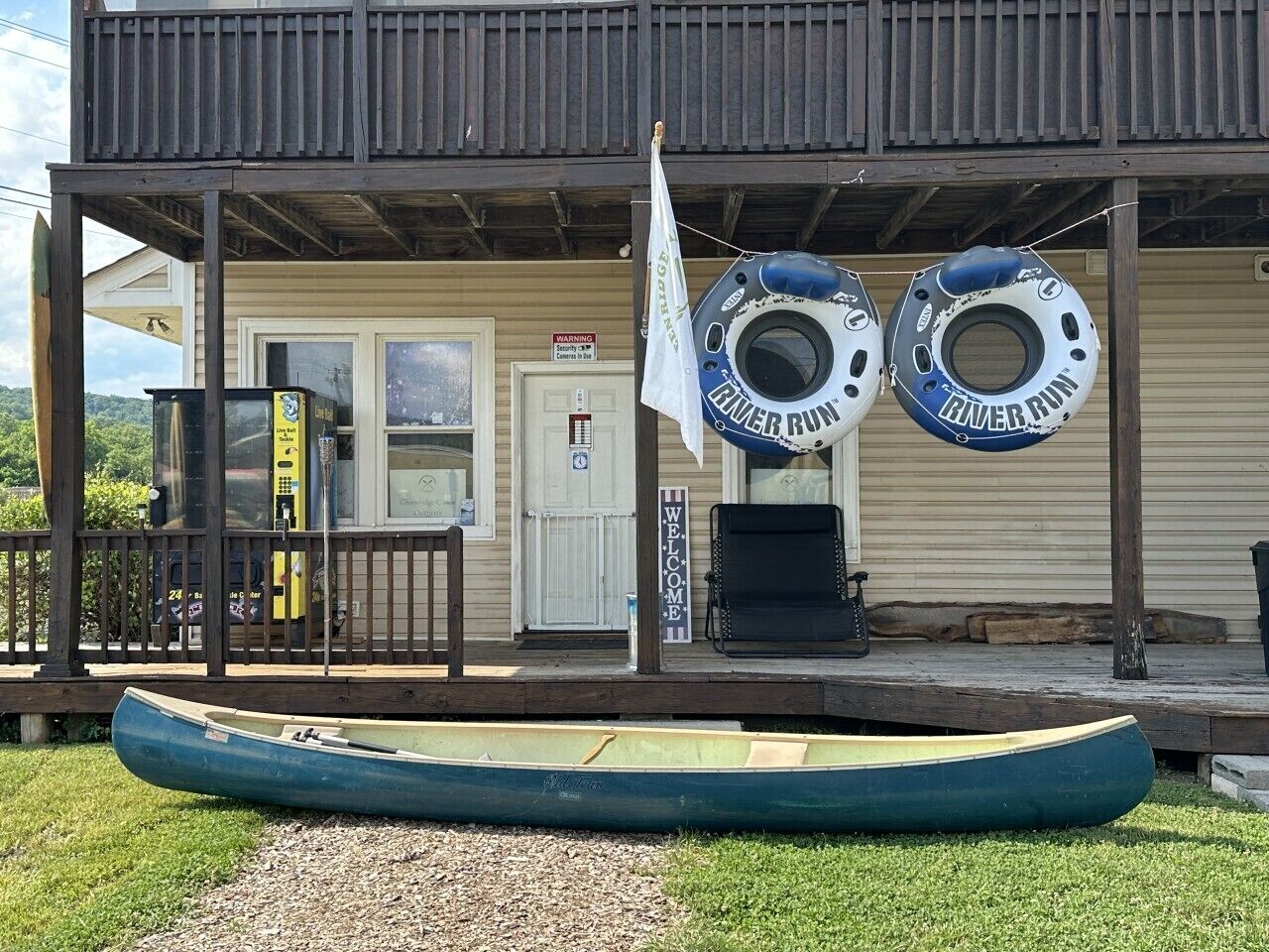 Un bâtiment doté d’un porche, avec deux bouées de sauvetage suspendues, une pancarte de bienvenue, une chaise et un canoë posé sur la pelouse.