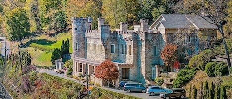 A castle-like building with stone walls and towers, surrounded by trees and a road with parked cars.