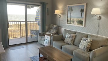 Relax in this inviting living room with a sofa set, coffee table, and wall-mounted lamps, offering a view of the beach through a sliding glass door.