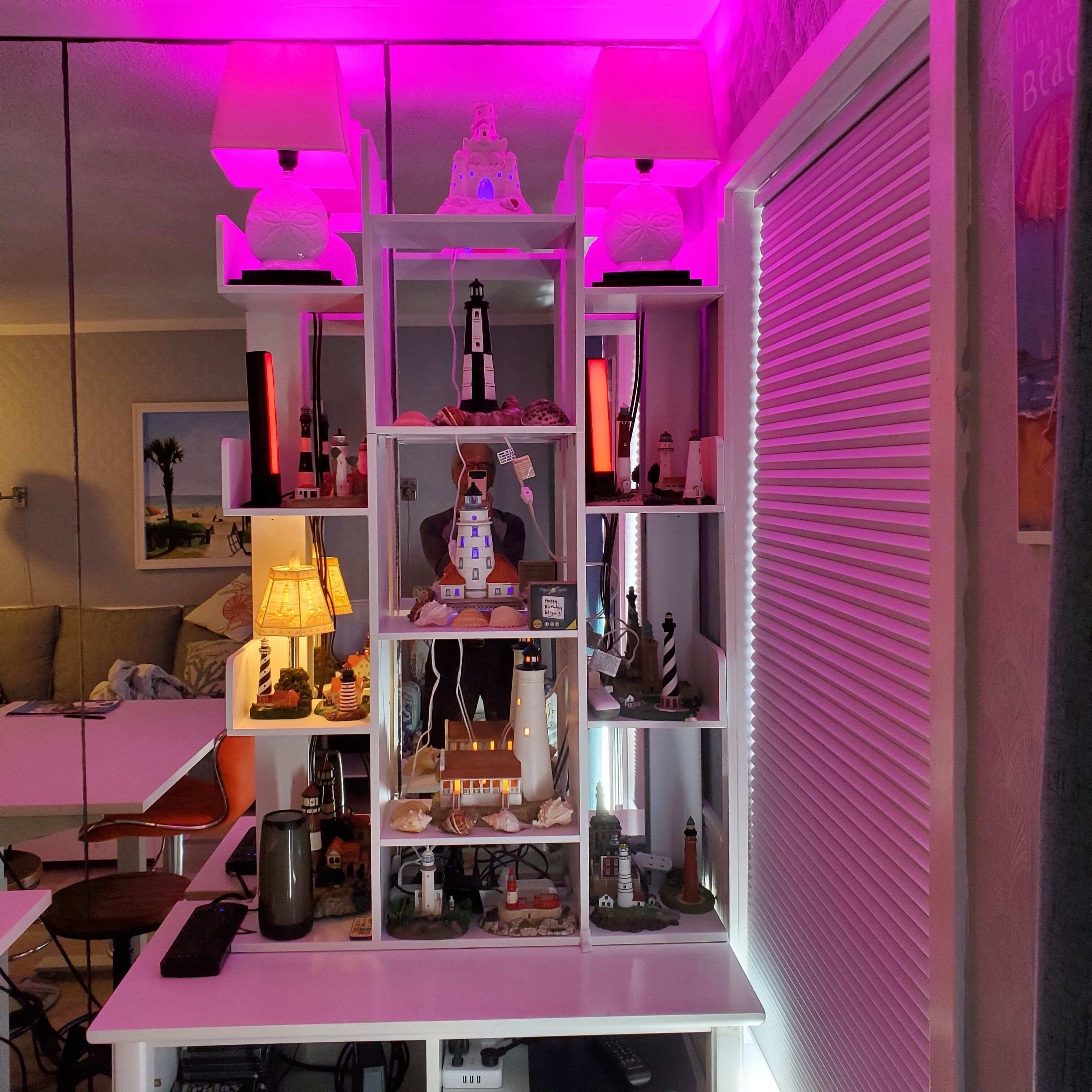 Een kamer met een wit bureau, planken met diverse spullen en een roze verlicht plafond.