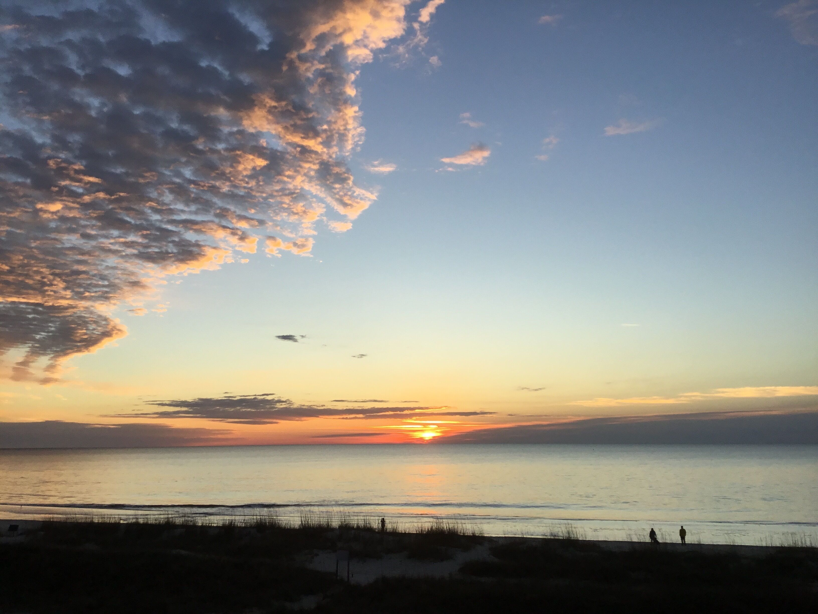 Escena de playa con un atardecer, mar tranquilo y siluetas de personas.