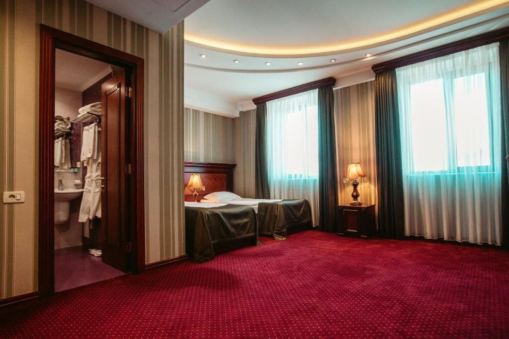 A hotel room with a red carpet, two beds, a wooden headboard, a nightstand with a lamp, and a bathroom visible through an open door.