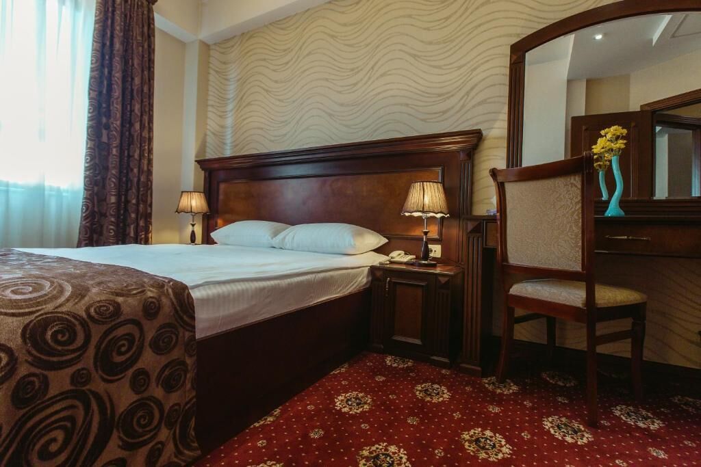 A hotel room with a bed, two bedside lamps, a chair, a mirror, and a patterned carpet.