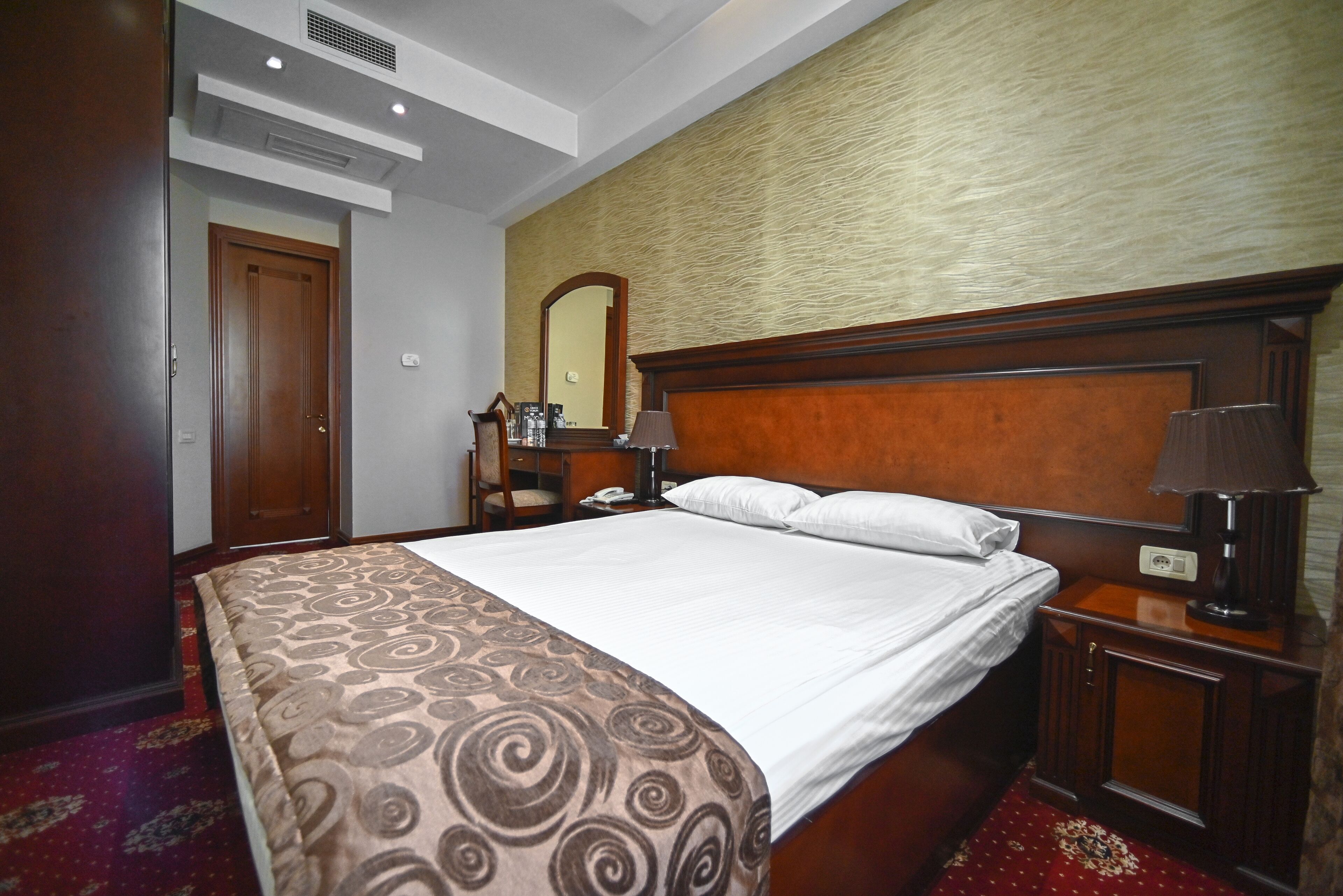 A hotel room with a large bed, wooden headboard, two bedside tables with lamps, a nightstand, a mirror, and a red carpeted floor.