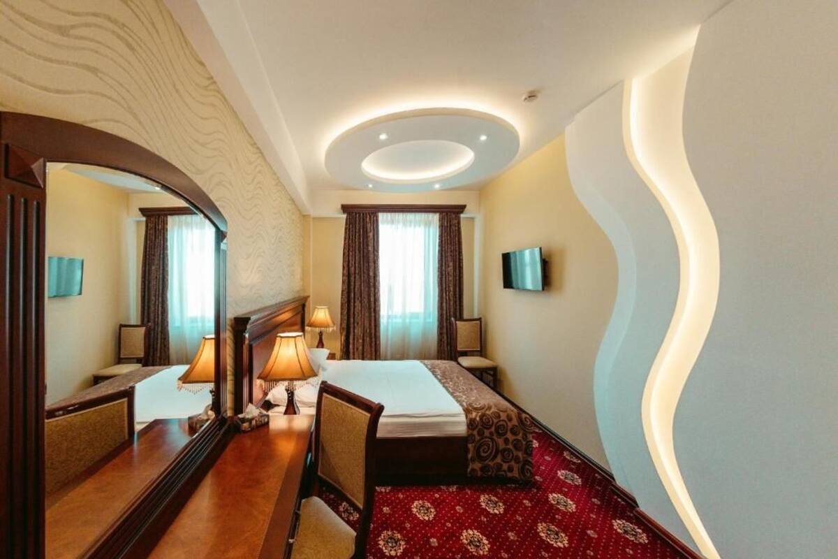 A hotel room with a bed, bedside tables, a desk, a mirror, a TV, and a unique ceiling design.