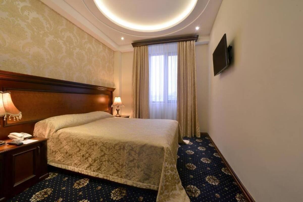 A hotel room with a large bed, two bedside lamps, a flat-screen TV mounted on the wall, and a patterned carpet.