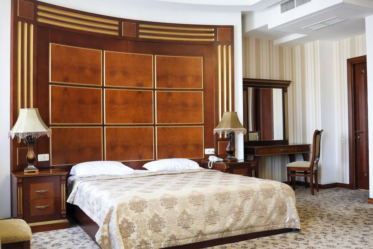 A hotel room with a large bed, a wooden headboard, a desk with a chair, a mirror, and a lamp.