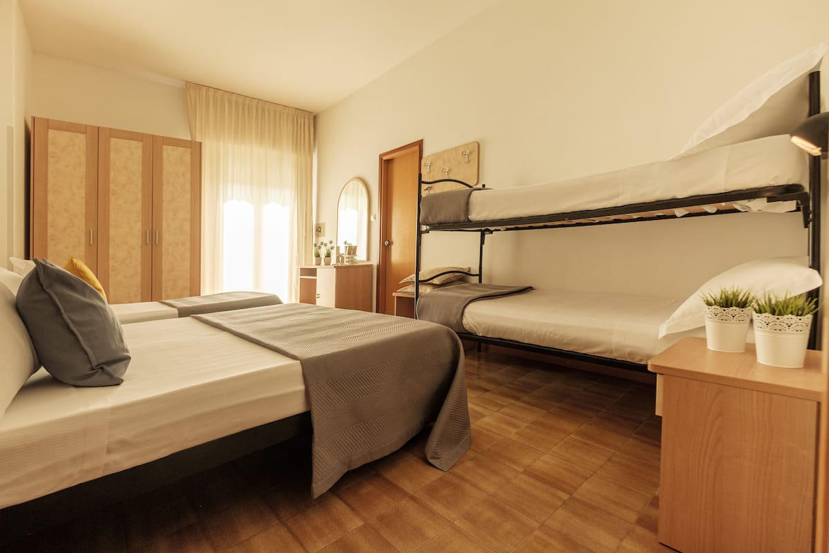 A hotel room with a bunk bed, a single bed, a wooden wardrobe, a bedside table with a lamp, and a mirror.