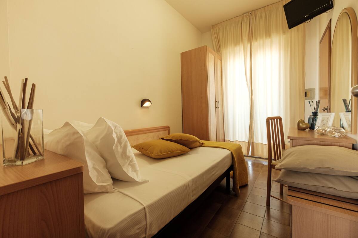 A hotel room with a bed, a wooden nightstand, a desk with a chair, a TV mounted on the wall, and a mirror.