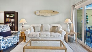 Un salon avec un canapé blanc, un fauteuil à motifs, une table basse et une planche de surf accrochée au mur.