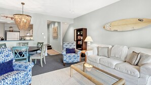 Un salon avec un canapé blanc, des fauteuils à motifs, une table basse, une bibliothèque et une planche de surf accrochée au mur.