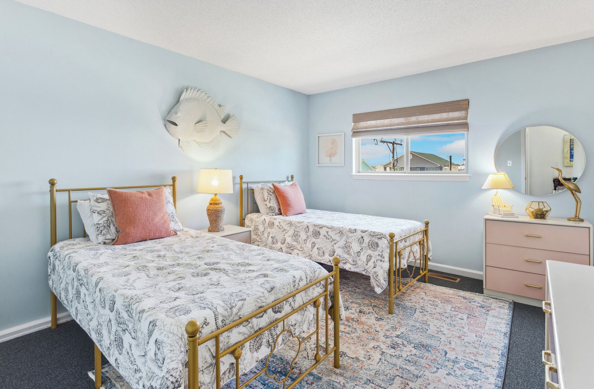 A bedroom with two beds, a dresser, a mirror, and a window with a view of houses.