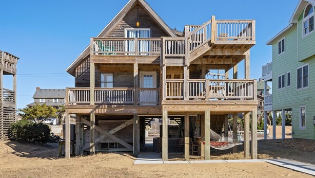 A multi-story wooden house with a balcony on each floor, a covered porch, and a sandy ground area. - 6129 Lady Jane * Semi-Oceanfront * Hot Tub (Nags Head)