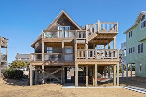 Une maison en bois à plusieurs étages, dotée d’un balcon à chaque niveau, d’un porche couvert et d’un espace au sol sablonneux.