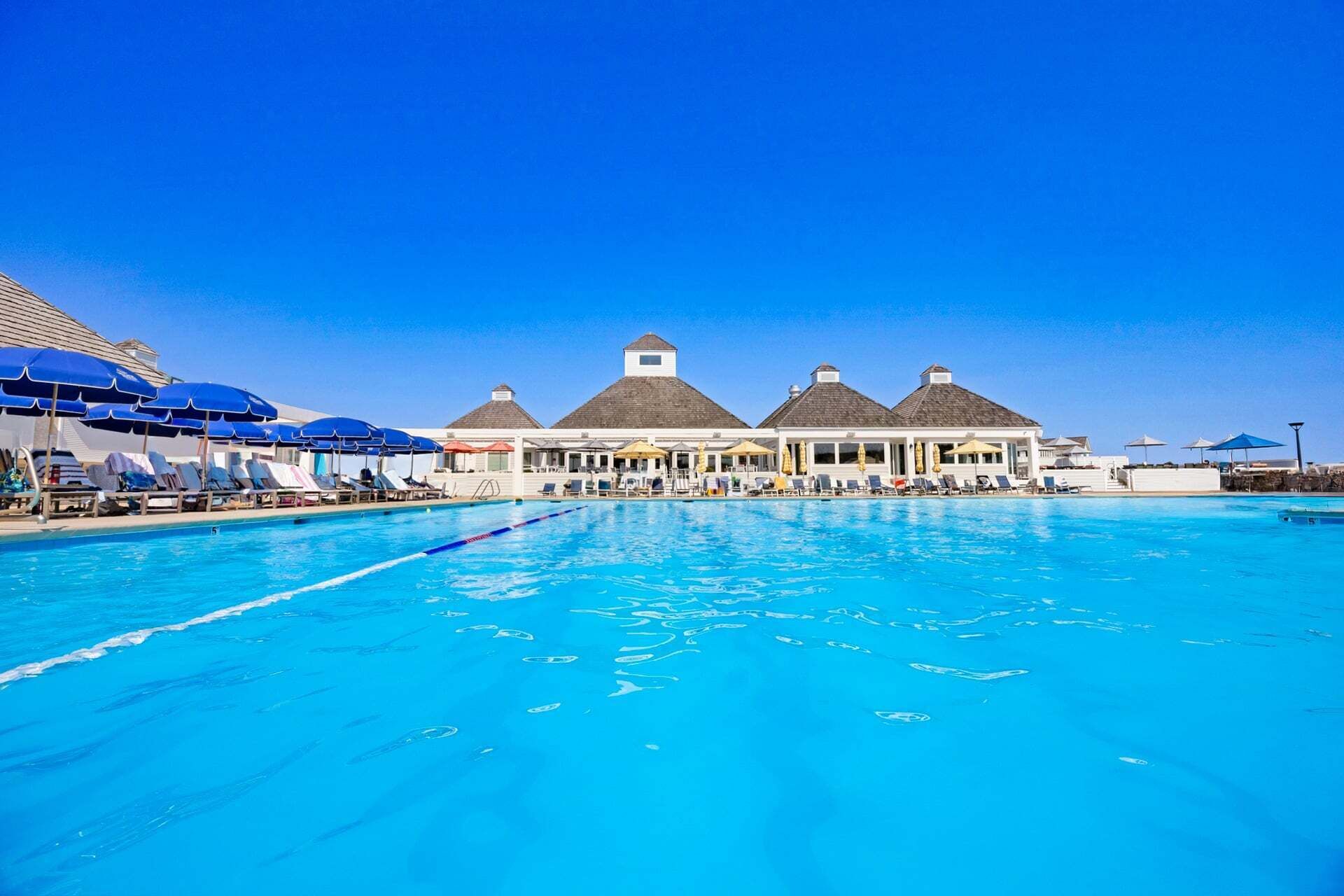 A large swimming pool with lounge chairs and umbrellas, surrounded by buildings with thatched roofs.
