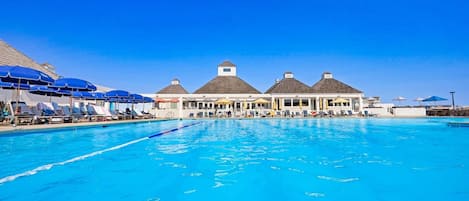 A large swimming pool with lounge chairs and umbrellas, surrounded by buildings with thatched roofs.