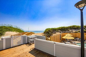 A wooden deck with a view of the beach, a pool area with umbrellas, and a white fence.