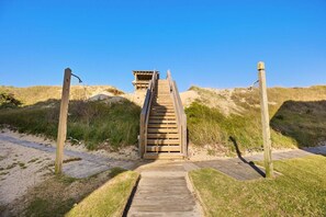 Una pasarela de madera que conduce a una casa de playa, con una playa arenosa y dunas al fondo.