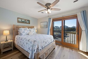 A bedroom with a bed, a nightstand, a ceiling fan, a window with curtains, and a painting on the wall.