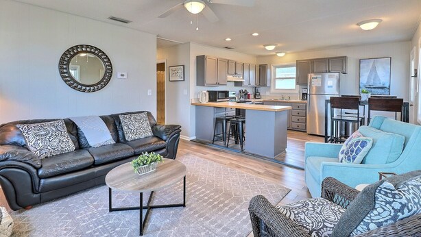 A modern living room with a black leather sofa, a round coffee table, and a blue armchair. A kitchen with a central island and bar stools is visible in the background. - High Five: Classic OBX Escape Near the Beach (Kill Devil Hills)