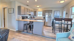 A modern kitchen with a central island, bar stools, and a dining area with a table and chairs. - High Five: Classic OBX Escape Near the Beach (Kill Devil Hills)