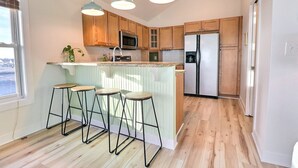 A modern kitchen with wooden cabinets, a stainless steel refrigerator, a microwave, and a bar area with stools. - The Shake Shack: Dog-Friendly + 450ft to Beach (Kitty Hawk)