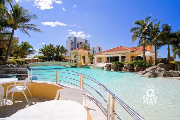 A resort pool area with clear blue water, palm trees, and a building with a red roof.