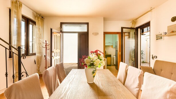 Un comedor con una mesa de madera, sillas color beige, un jarrón con flores y vista a la cocina a través de un umbral abierto.