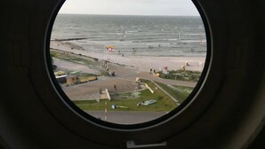 Vista a través de un ventanal circular de una playa con gente nadando e un asta con una bandera.