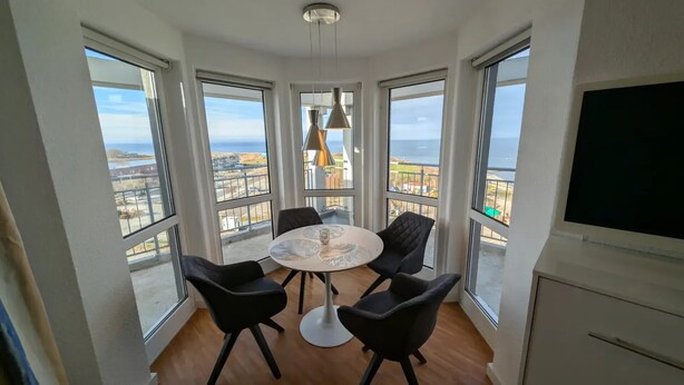 Habitación de esquina con una mesa redonda, cuatro sillas, un televisor y ventanas amplias con vistas.