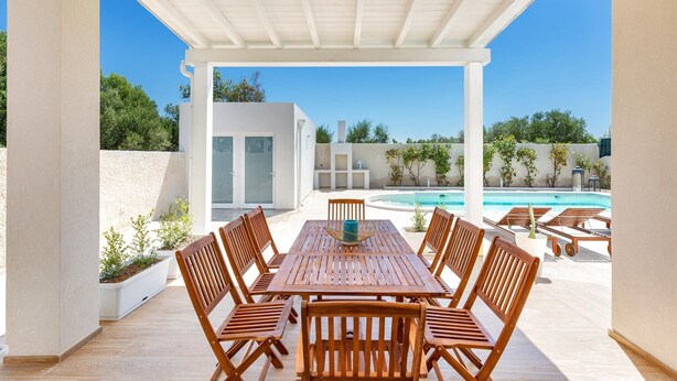 An outdoor patio with a wooden table and chairs, a pool in the background, and a white building. - 2055 Villa San Teodoro (Avetrana)