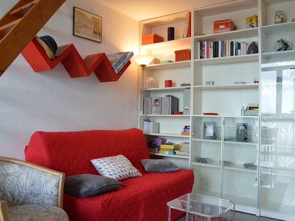 A living room with a red sofa, a patterned armchair, and a glass coffee table. There is a bookshelf filled with books and decorative items.