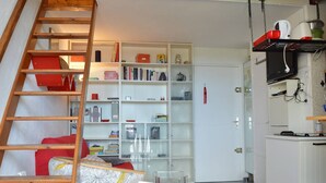 A compact living space with a wooden ladder leading to a loft area, a white bookshelf filled with books and decorative items, and a kitchen area with a microwave and a small refrigerator.