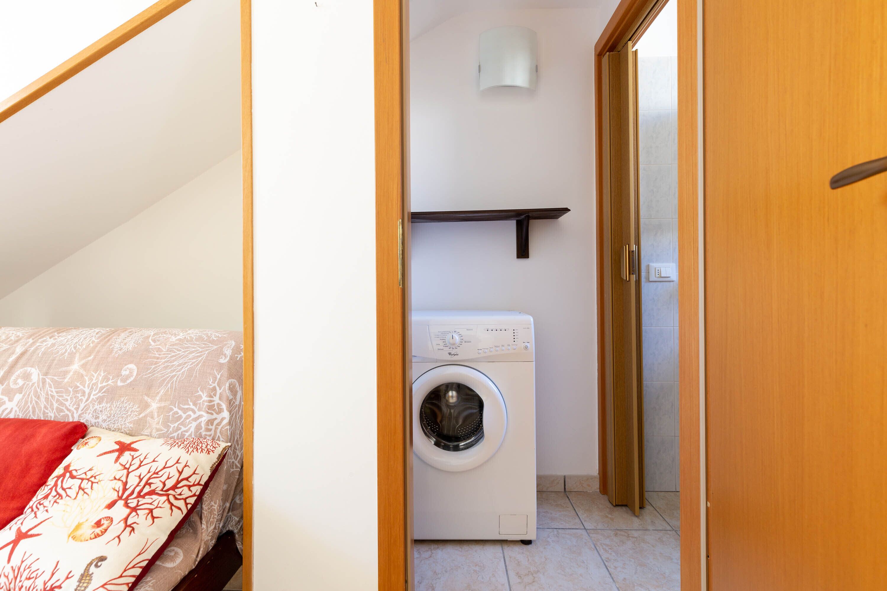 Une pièce de taille modeste, équipée d’un lave-linge, d’un lit recouvert d’une courtepointe à motifs et d’une porte en bois.