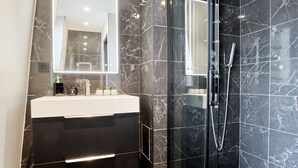 A modern bathroom with a marble-tiled shower, a white sink, and a mirror above it. - Blackpearl-HyperKing Bed CenterSize-Parking Offered (Amiens)