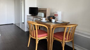 Ein kleiner, sauberer Raum mit Schreibtisch, zwei Korbstühlen, einem Fernseher, einem Korb, einer weißen Tasse und einem gerahmten Bild an der Wand.