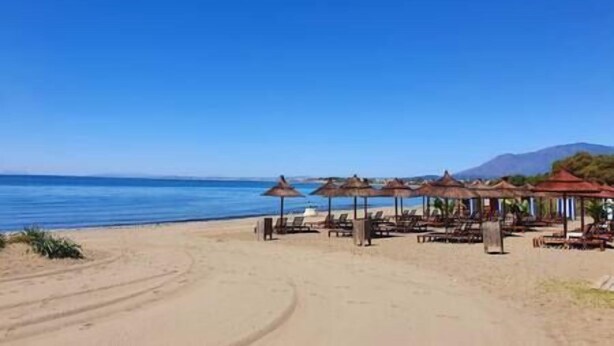 A beach with sun umbrellas and lounge chairs, a clear sky, and a mountain in the background.