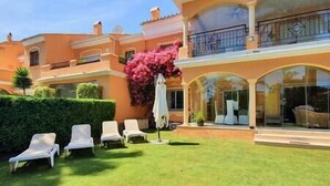 A large house with a well-maintained lawn, white lounge chairs, and a pink flowering tree.
