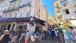 A bustling street with outdoor seating at cafes, a bakery, and a market stall selling postcards.