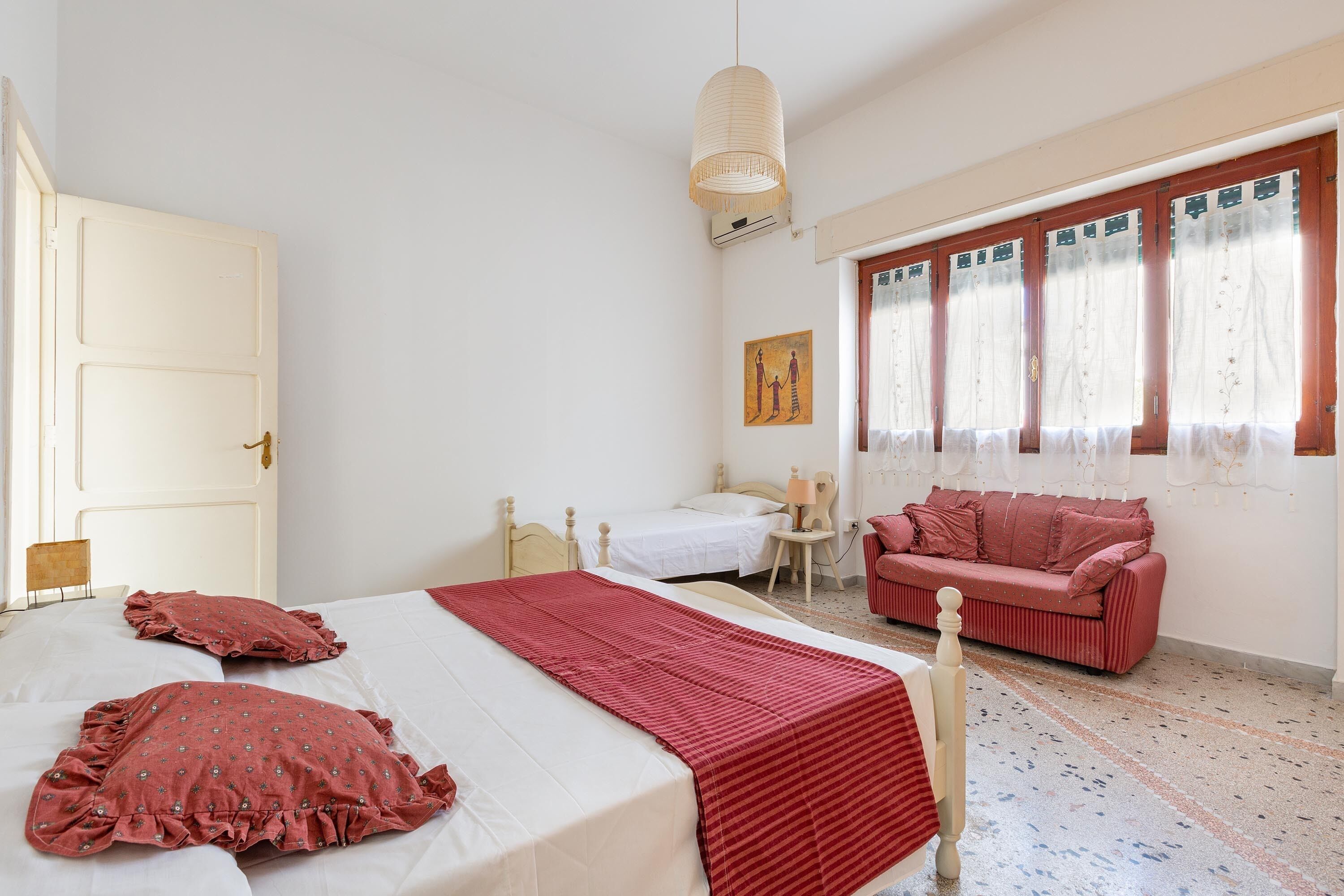 A bedroom with a bed, a red sofa, a small table, and a window with curtains.