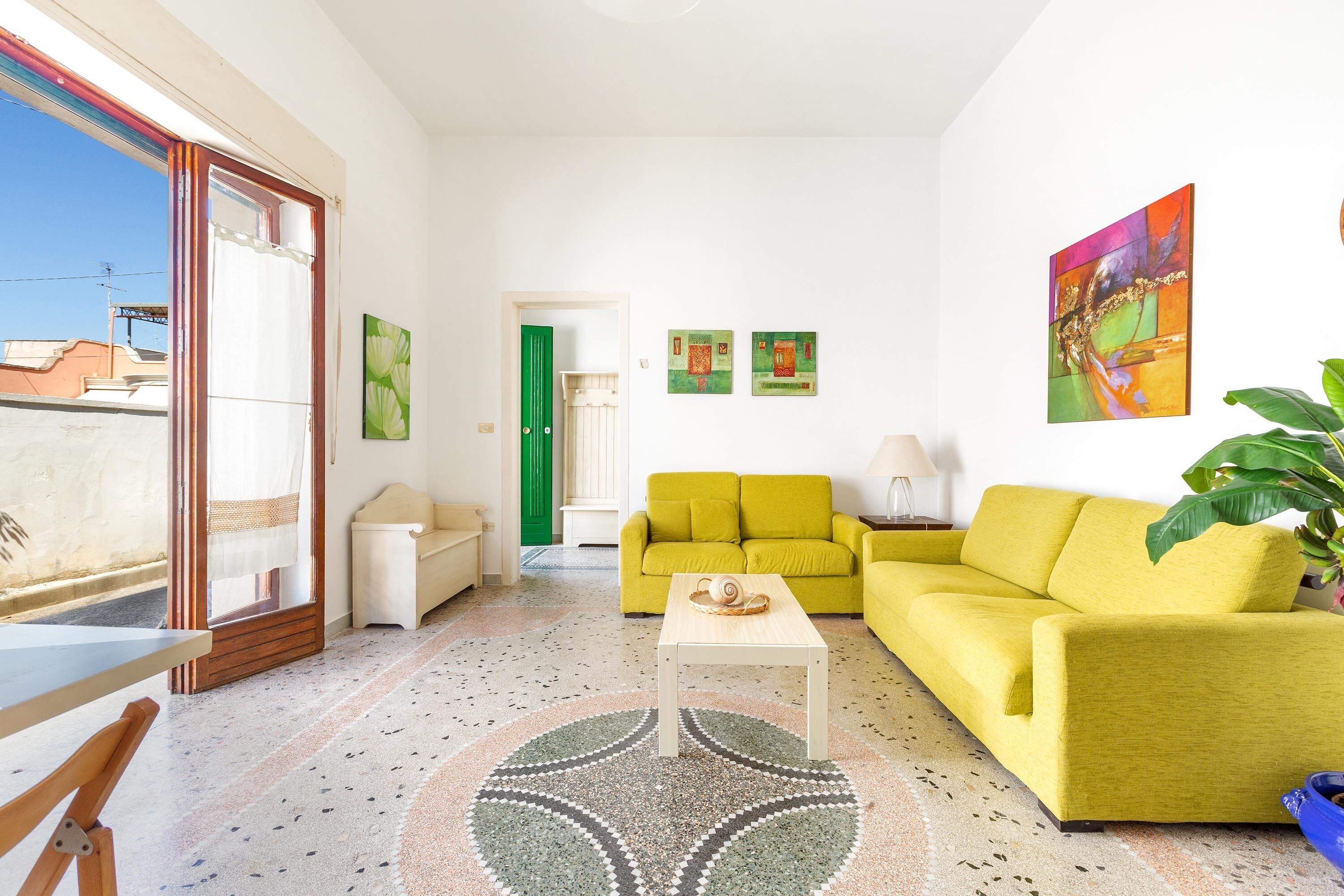 A living room with a yellow sofa, a small table, and a green door. There are three paintings on the wall and a plant in the corner.