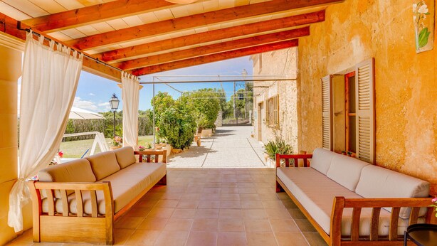Eine überdachte Terrasse mit Holzbanken und einer Couch, Blick auf einen Garten sowie eine rustikale Wand.