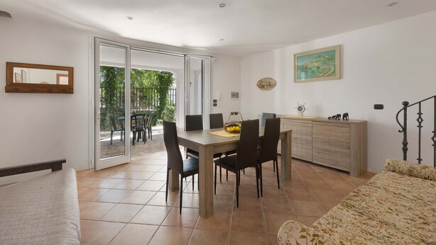 Une pièce comprenant un lit, une table à manger avec des chaises, un meuble et une porte-fenêtre coulissante donnant sur un espace extérieur aménagé avec des chaises et de la verdure.
