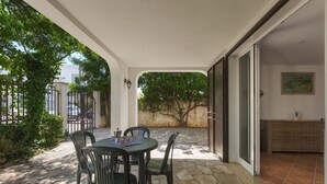Une terrasse extérieure avec une table et des chaises, un sol en pierre, une vue sur un arbre et l’entrée d’un bâtiment.