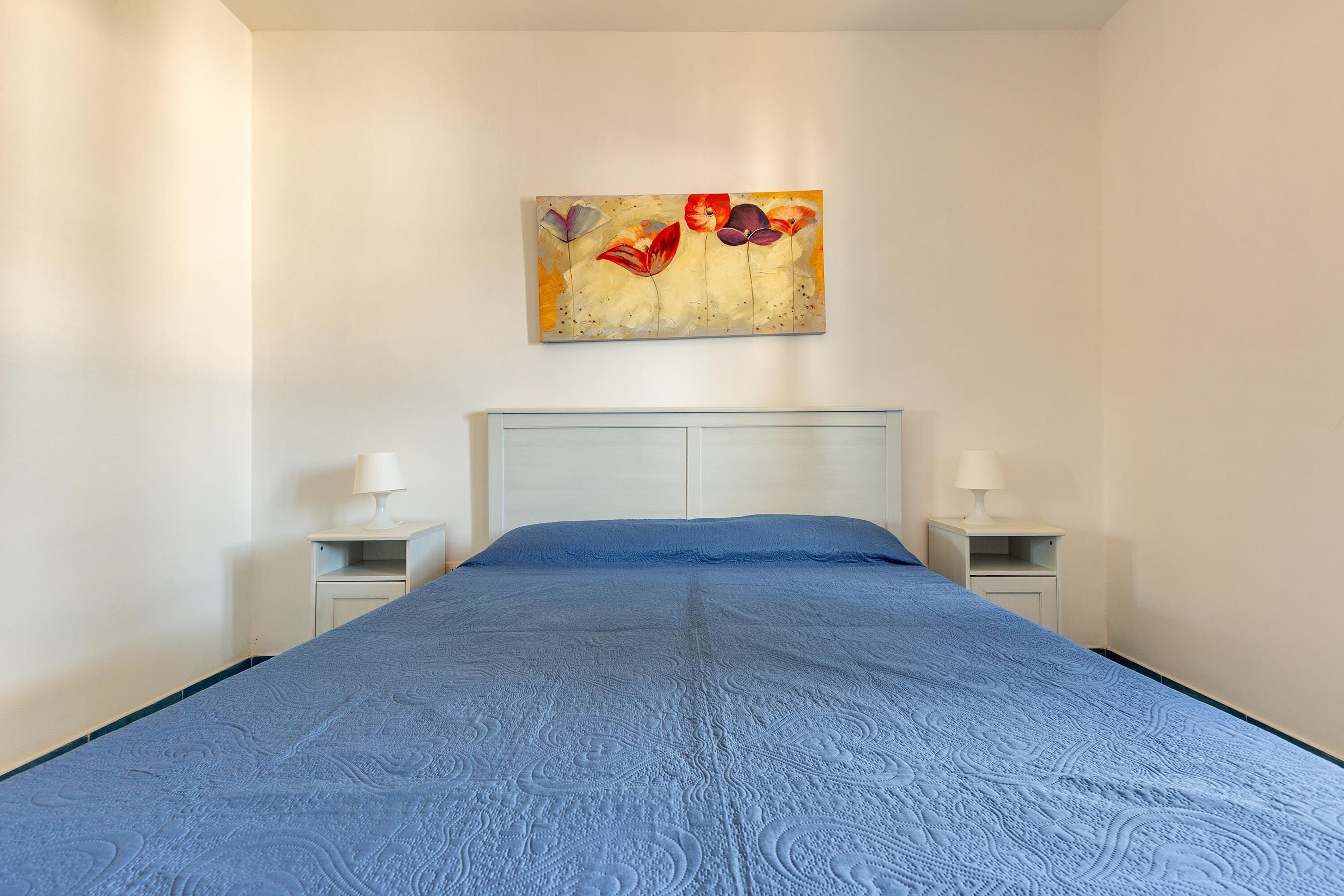 A bedroom with a large bed, white headboard, two bedside tables with lamps, and a wall-mounted painting.