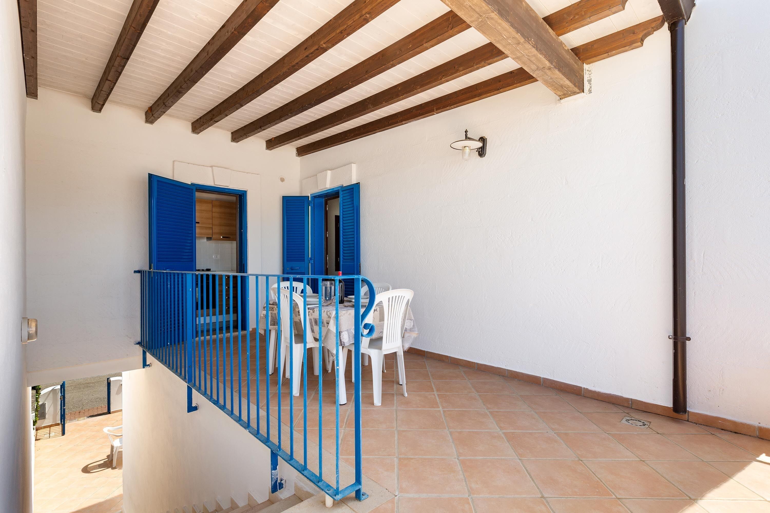 A terrace with a table and chairs, a blue railing, and a white wall with a door.